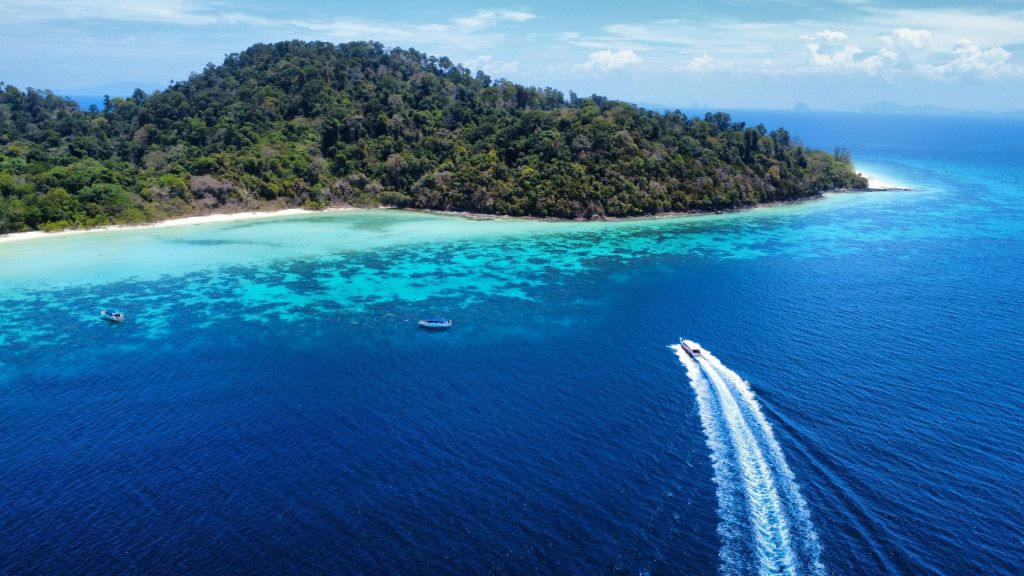 fotka z drona na národný park, zelený neobývaný ostrov, tmavomodé more a koraly. Loď plaviaca sa na ostov zanecháva stopu v mori