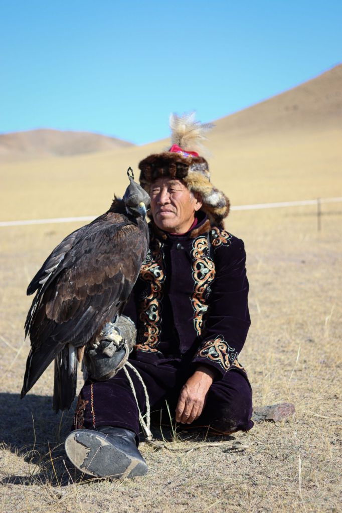 mongol v tradičnom vyšívanom oblečení v čiapke s kožušinou pozerajúci sa na orla ktorého drží v ruke